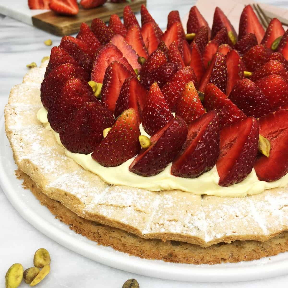 Pistachio dacquoise decorated with fresh strawberries on a white serving platter with berries and flatware on background.