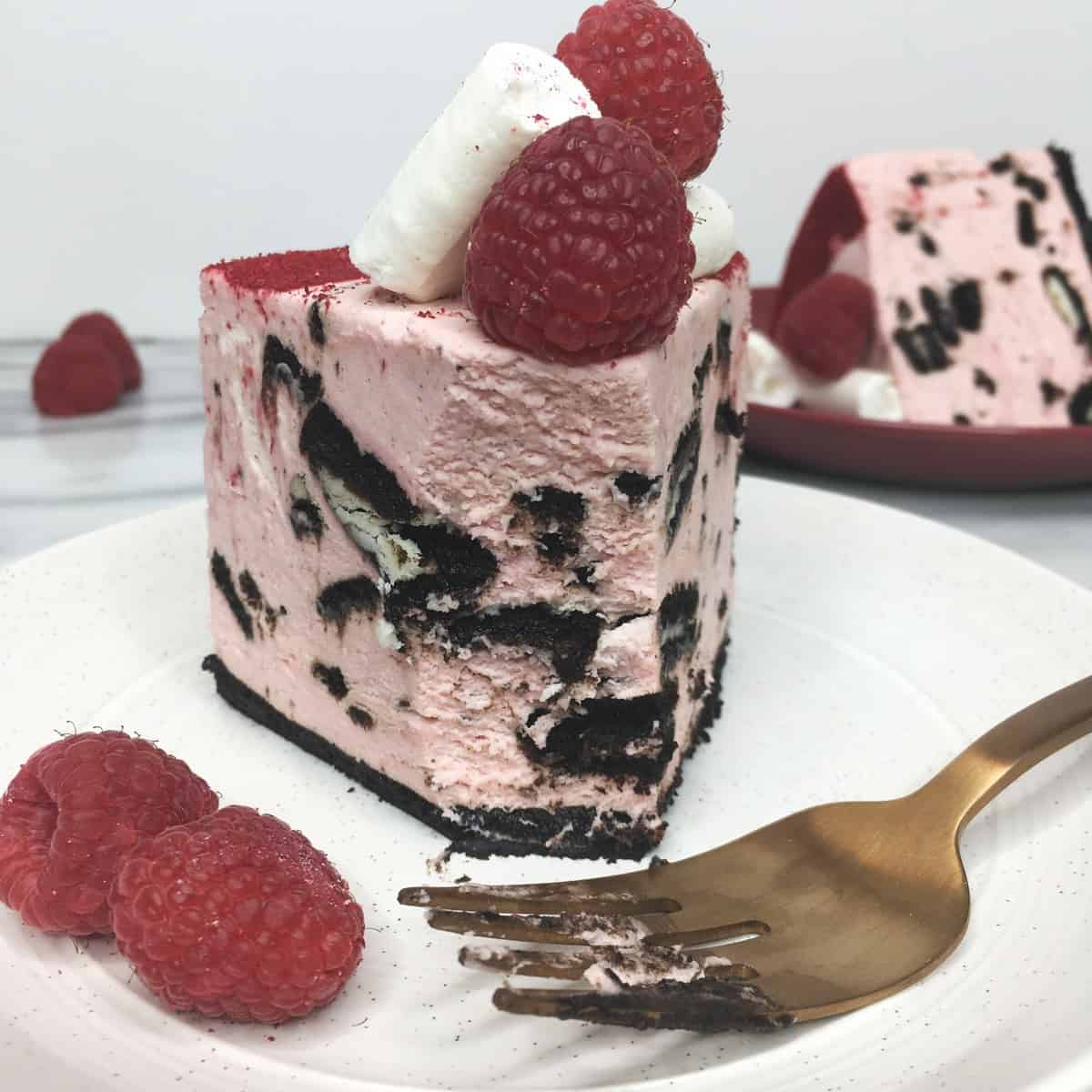 A slice of raspberry cheesecake with Oreo cookie crust, a fork, raspberries on a white plate 