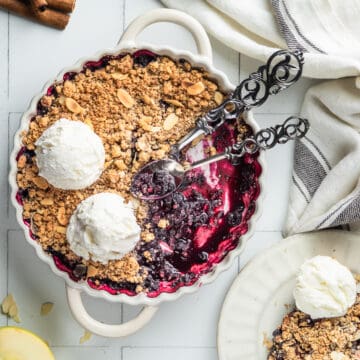 Apple and blueberry crumble in a white ceramic dish with ice cream and two spoons.